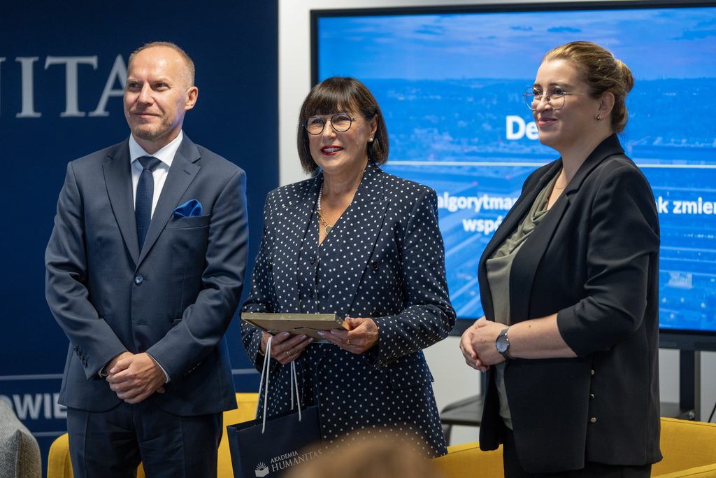 Przedstawiciele Akademii Humanitas pozują z uczestniczką wydarzenia, która trzyma pamiątkowy akt powołania i torbę upominkową. W tle widoczny jest ekran z grafiką debaty oraz elementy identyfikacji wizualnej uczelni.