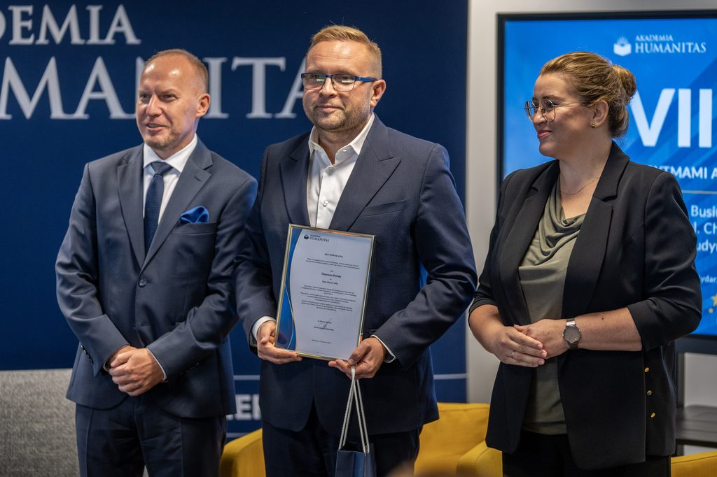 Przedstawiciele Akademii Humanitas pozują z uczestnikiem wydarzenia, który trzyma pamiątkowy akt powołania i torbę upominkową. W tle widoczny jest ekran z grafiką debaty oraz elementy identyfikacji wizualnej uczelni.