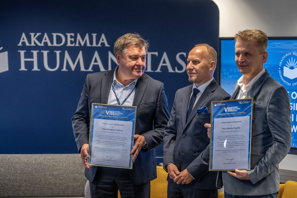 Przedstawiciele Akademii Humanitas pozują z partnerami wydarzenia, którzy trzymają pamiątkowe podziękowania VIII Kongresu Praktyków Biznesu. W tle widoczna jest granatowa ścianka z logotypem uczelni.