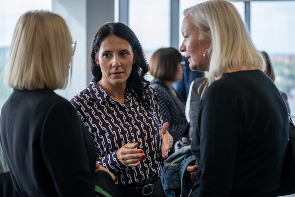 Uczestniczki wydarzenia rozmawiają podczas przerwy networkingowej w przestrzeni konferencyjnej. W tle widoczne są panoramiczne okna oraz inni uczestnicy spotkania.