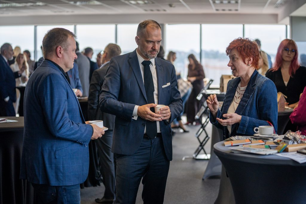 Uczestnicy wydarzenia rozmawiają przy stoliku podczas przerwy kawowej. W tle widoczna jest przestrzeń networkingowa z innymi gośćmi spotkania.