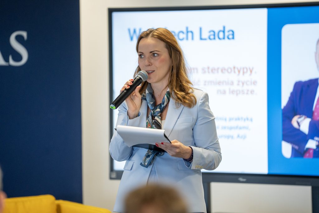 Prowadząca wydarzenie przemawia do mikrofonu, trzymając notatki. W tle widoczny jest ekran z prezentacją prelegenta oraz elementy identyfikacji wizualnej Akademii Humanitas.
