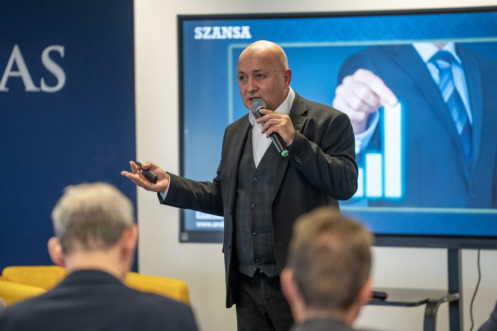 Prelegent omawia prezentację podczas wydarzenia biznesowego Akademii Humanitas. Mężczyzna przemawia z mikrofonem, a na ekranie za nim widoczny jest slajd z hasłem „Szansa”.