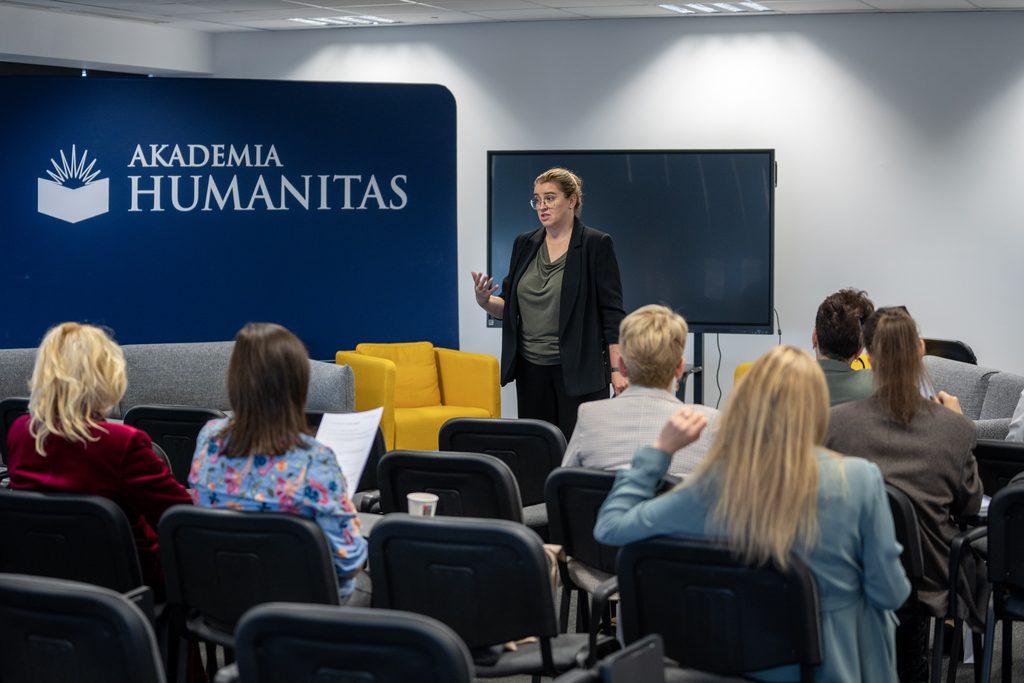 Przedstawicielka Akademii Humanitas prowadzi wystąpienie przed publicznością w sali konferencyjnej. W tle widoczna jest granatowa ścianka z logotypem uczelni oraz ekran prezentacyjny.