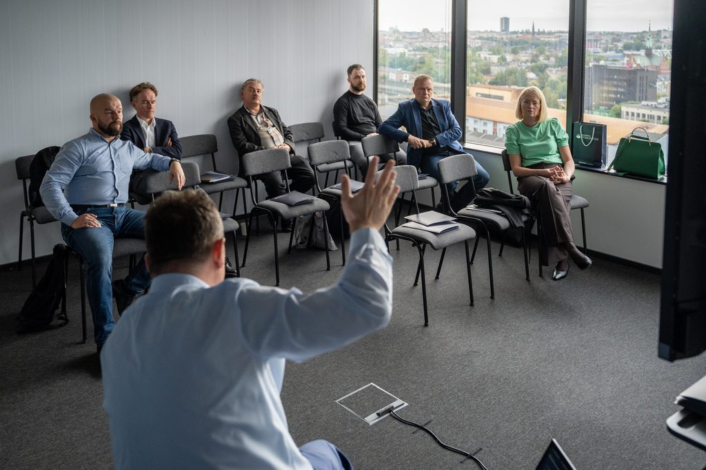 Uczestnicy szkolenia siedzą w sali z panoramicznymi oknami i słuchają prowadzącego. Spotkanie odbywa się w kameralnej przestrzeni konferencyjnej z widokiem na miasto.