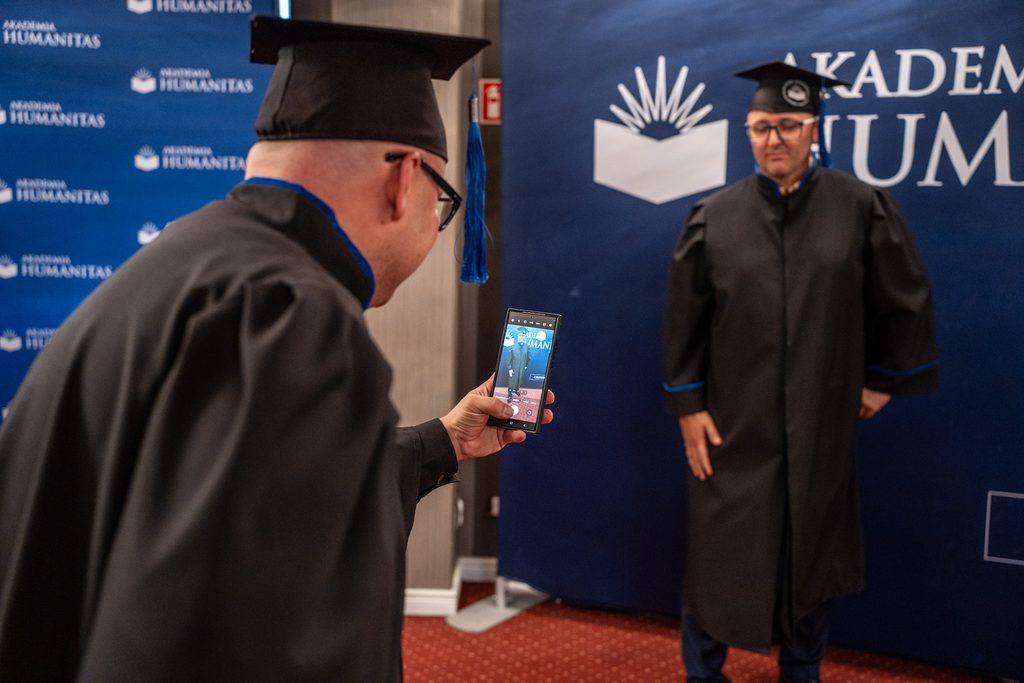 Osoba w todze wykonuje zdjęcie pamiątkowe telefonem drugiej osobie stojącej przy ściance Akademii Humanitas.