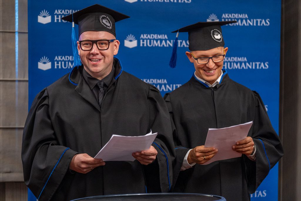 Dwóch absolwentów Akademii Humanitas w togach i biretach odczytuje tekst podczas uroczystości.