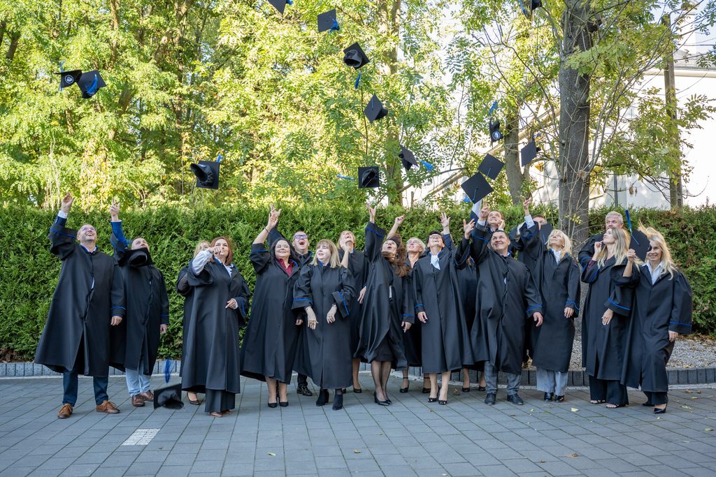 Absolwenci Akademii Humanitas podrzucają birety w górę podczas pamiątkowego zdjęcia plenerowego.