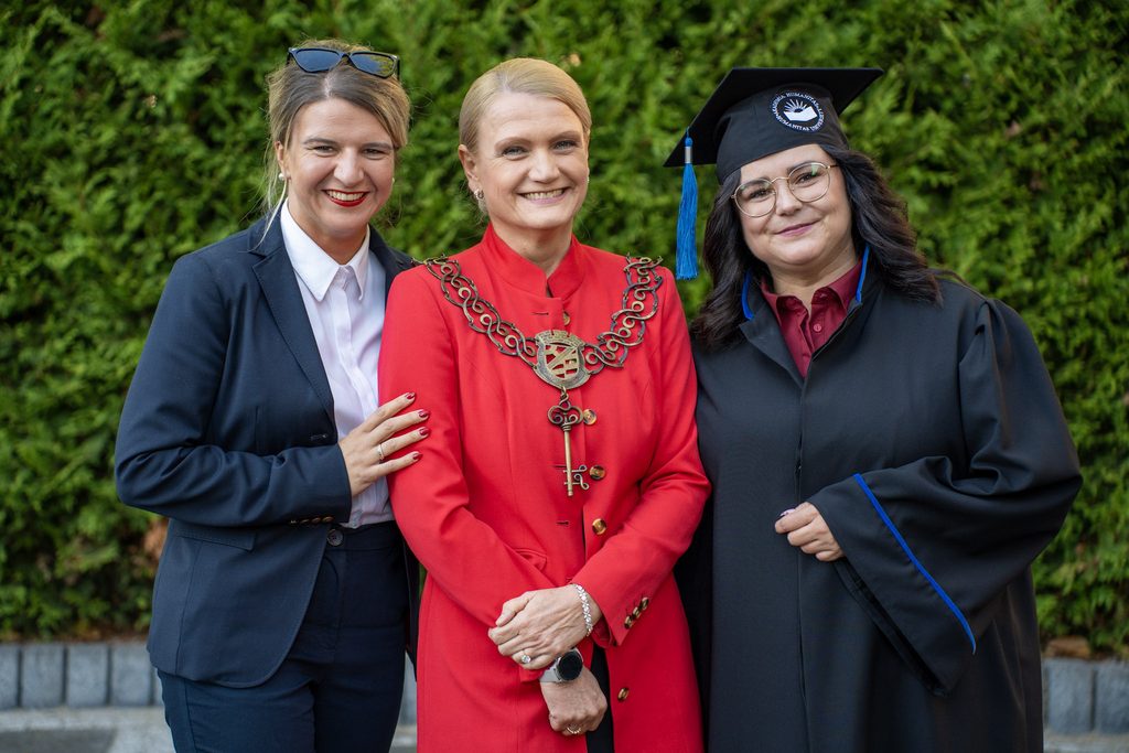 Trzy kobiety pozują do pamiątkowego zdjęcia podczas uroczystości akademickiej, jedna z nich ma na sobie togę i biret.