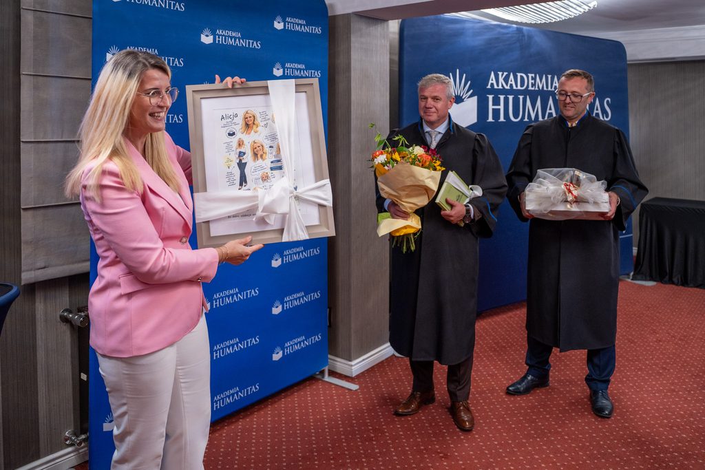 Kobieta w różowym garniturze prezentuje oprawiony pamiątkowy upominek podczas uroczystości Akademii Humanitas.