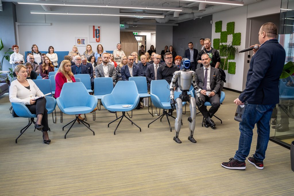 Uczestnicy wydarzenia obserwują prezentację humanoidalnego robota, a prowadzący omawia jego możliwości z mikrofonem w dłoni.