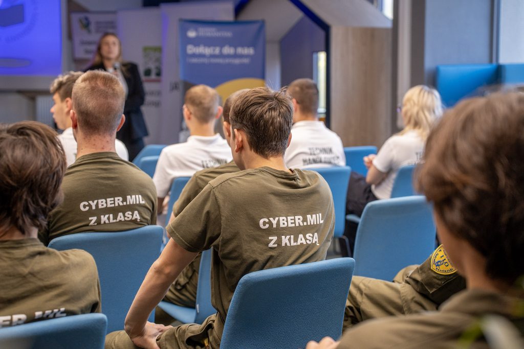 Uczestnicy w koszulkach z napisem „Cyber.Mil z klasą” słuchają prezentacji przedstawicielki Akademii Humanitas.