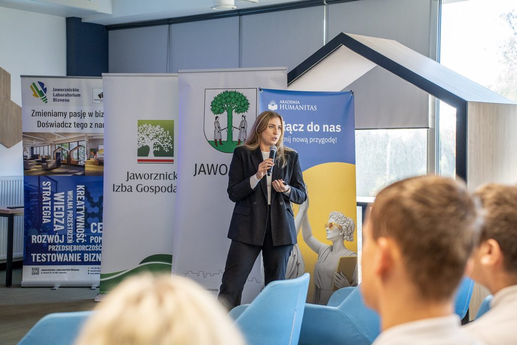 Przedstawicielka Akademii Humanitas przemawia do uczestników wydarzenia na tle banerów uczelni oraz jaworznickich instytucji.