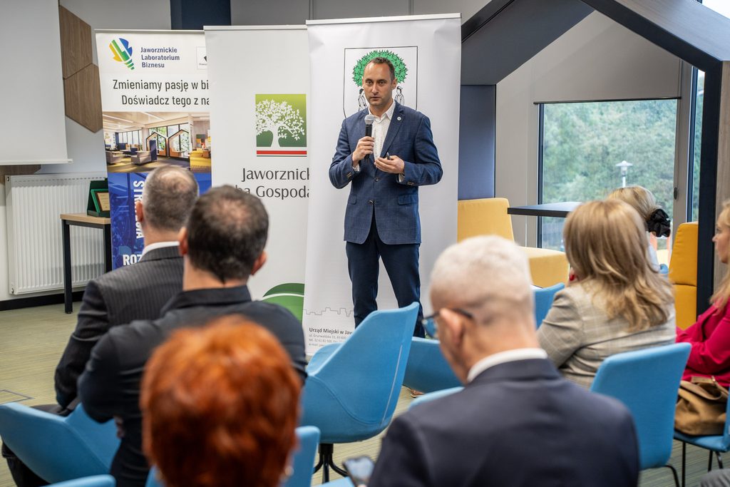 Mężczyzna w niebieskiej marynarce przemawia do uczestników wydarzenia na tle banerów Jaworznickiego Laboratorium Biznesu i Jaworznickiej Izby Gospodarczej.