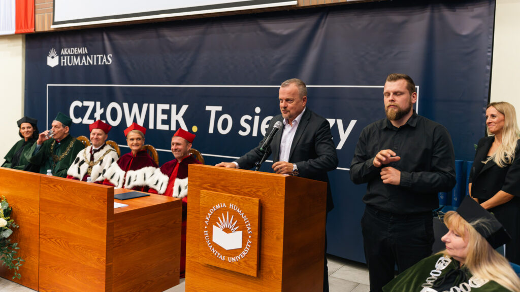 Prezydent Miasta Sosnowiec Arkadiusz Chęciński przemawia przy mównicy, a obok tłumacz języka migowego przekłada jego wystąpienie. W tle władze Uczelni w togach akademickich.