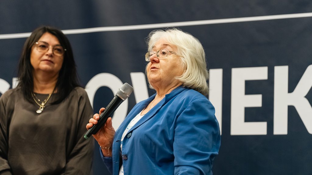 Kobieta w niebieskiej marynarce przemawia do uczestników wydarzenia, trzymając mikrofon, na tle hasła Akademii Humanitas.