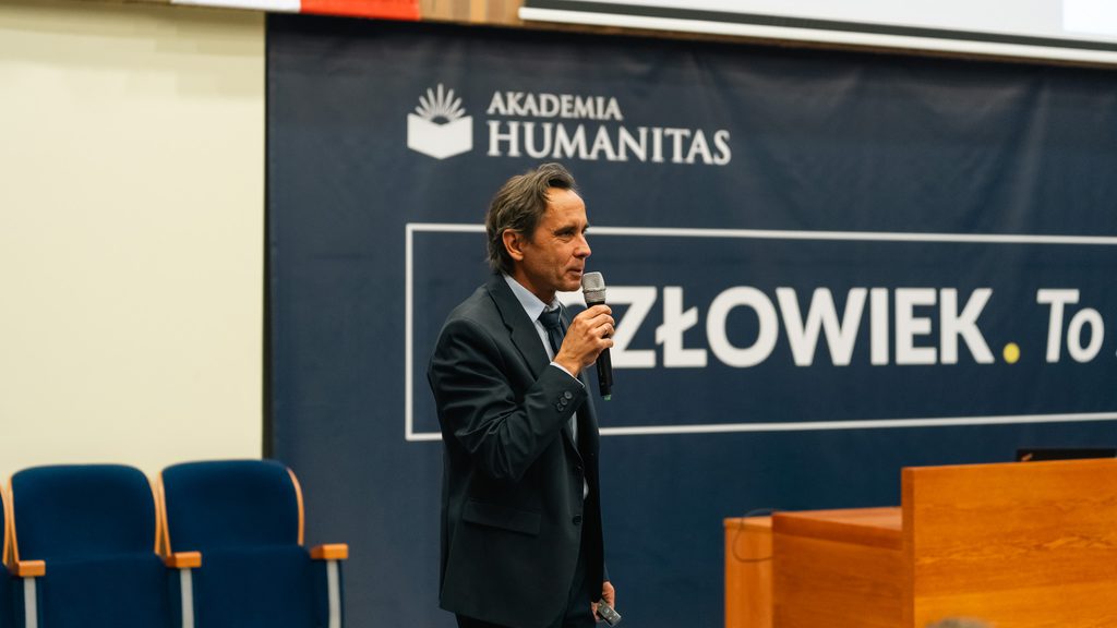 Prelegent prowadzi wystąpienie z mikrofonem na tle ścianki Akademii Humanitas z hasłem „Człowiek. To się liczy”.