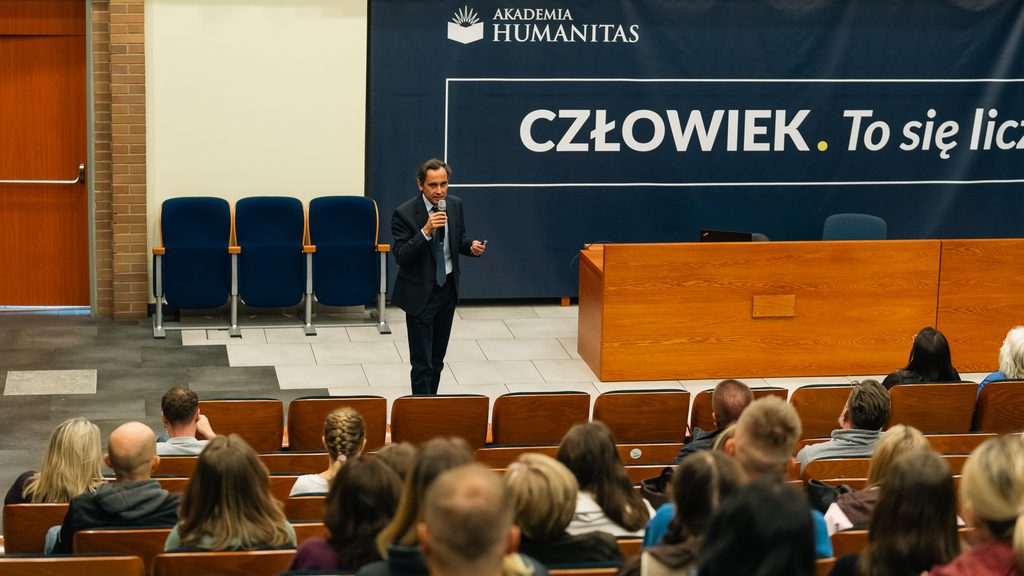 Prowadzący z mikrofonem wygłasza wykład w auli Akademii Humanitas przed zgromadzoną publicznością.