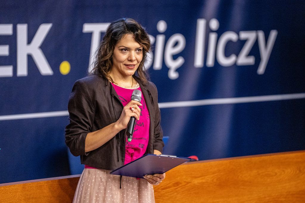 Prelegentka Girls’ Week przemawia z mikrofonem na tle hasła "Człowiek. To się liczy". 