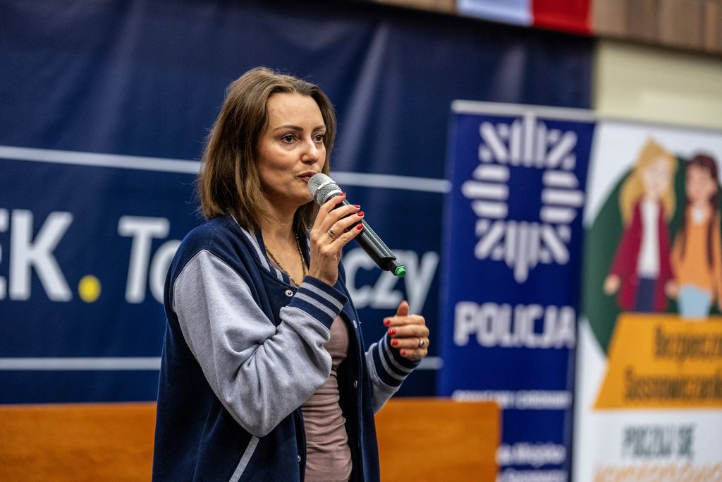Prelegentka z mikrofonem prowadzi wystąpienie podczas Girls’ Week na tle baneru Policji.