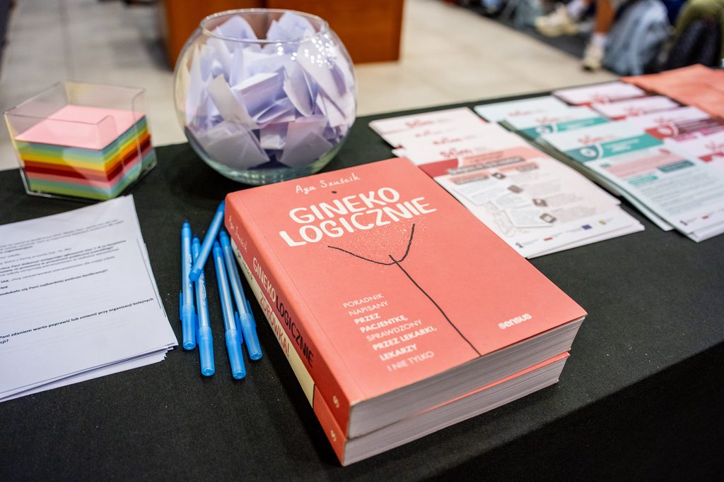 Na stoliku widoczne są materiały edukacyjne, ankiety, kolorowe karteczki oraz książka „Gineko logicznie”.