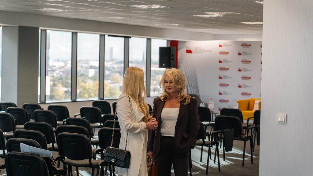 Dwie uczestniczki wydarzenia rozmawiają w sali konferencyjnej przed rozpoczęciem spotkania.