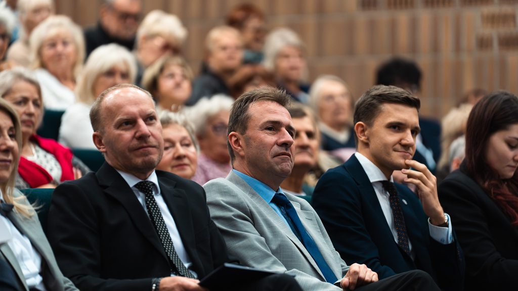 Trzech mężczyzn w eleganckich garniturach siedzi w pierwszym rzędzie auli podczas inauguracji Uniwersytetu Trzeciego Wieku.