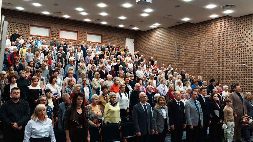 Uczestnicy inauguracji Uniwersytetu Trzeciego Wieku stoją w wypełnionej auli Akademii Humanitas podczas uroczystej części wydarzenia.