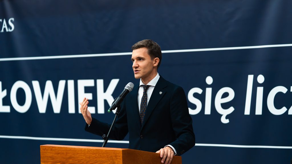 Mężczyzna w granatowym garniturze przemawia do uczestników inauguracji Uniwersytetu Trzeciego Wieku.