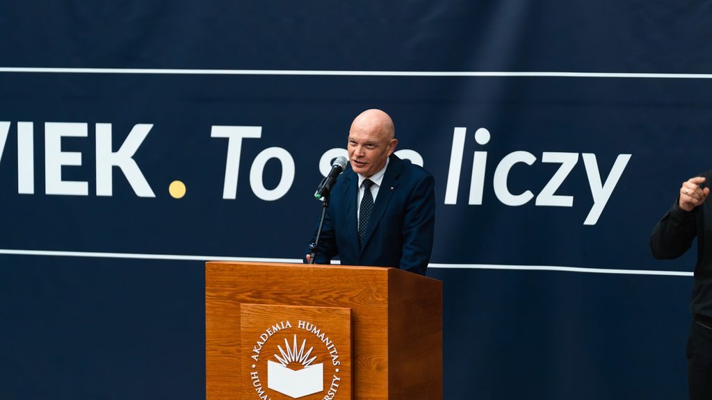 Mężczyzna w granatowym garniturze przemawia zza drewnianej mównicy Akademii Humanitas. Z prawej strony widoczny fragment tłumacza języka migowego.