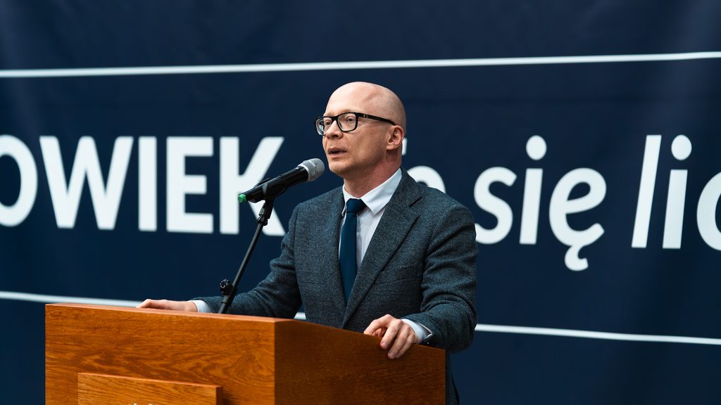 Mężczyzna w okularach, szarym garniturze i granatowym krawacie przemawia przy mównicy podczas inauguracji Uniwersytetu Trzeciego Wieku.