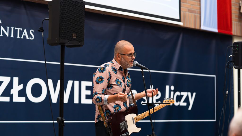 Muzyk w różowej koszuli w kwiaty stoi przy mikrofonie z gitarą elektryczną podczas występu artystycznego na inauguracji UTW.