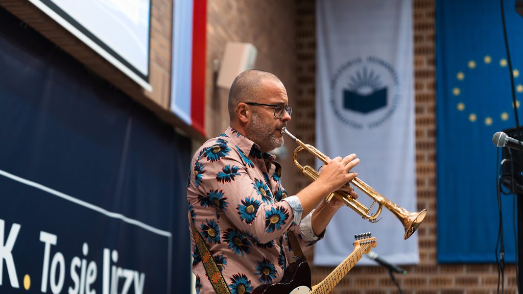 Muzyk w różowej koszuli w kwiaty gra na trąbce podczas występu w auli Akademii Humanitas. Na ramieniu ma zawieszoną gitarę elektryczną.