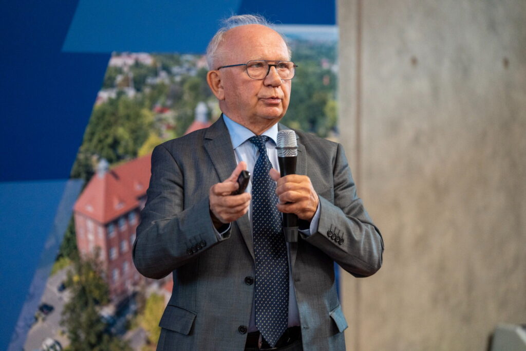 Profesor Krystian Roleder wygłasza wykład inauguracyjny, trzymając mikrofon i pilot do prezentacji multimedialnej.